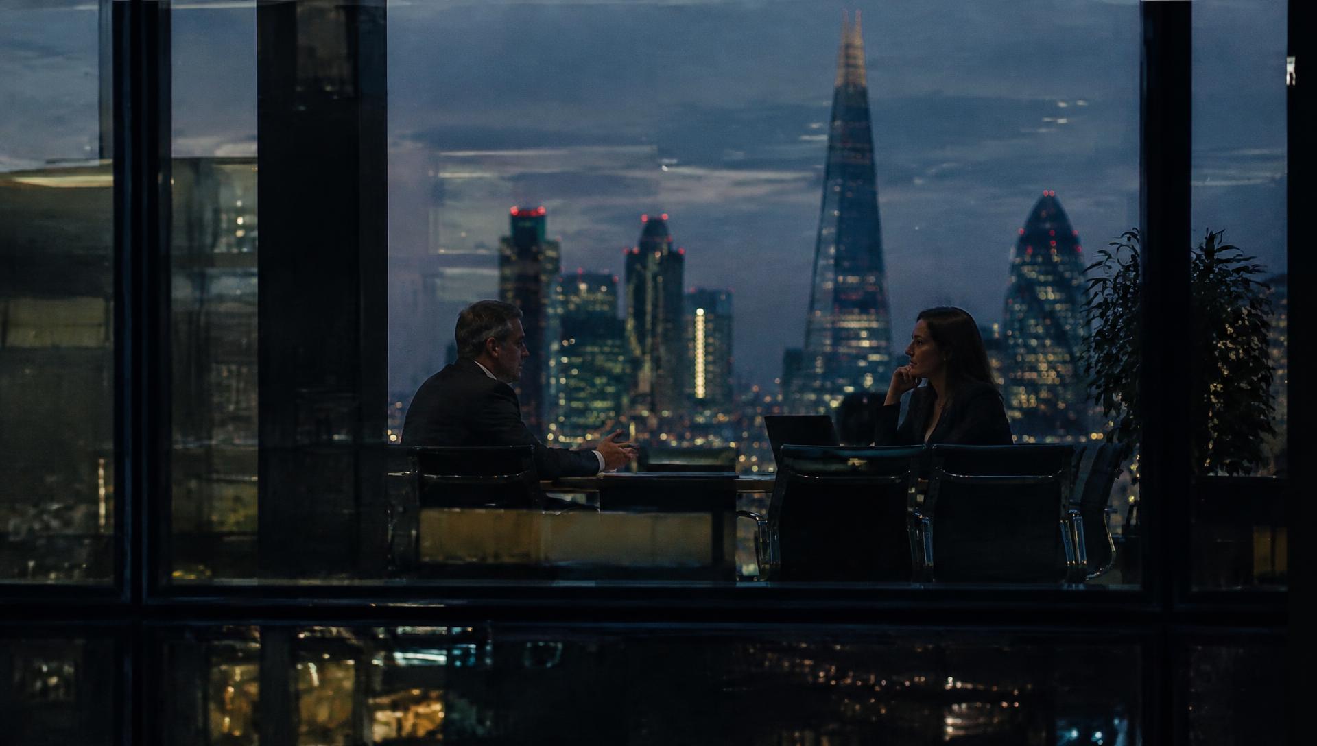 Two business people in conversation at a meeting table, seen through an office window at dusk with the London skyline behind